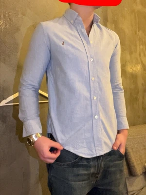 Ljusblå skjorta från Ralph Lauren - Klassisk ljusblå skjorta från Ralph Lauren med broderad logga på bröstet. Skjortan har knappar hela vägen, button-down-krage och lång ärm. Tillverkad i mjuk bomull och har en clean, normal passform som funkar till allt.