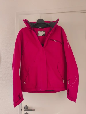 Rosa vindjacka från Salomon M - Säljer en snygg rosa vindjacka från Salomon i storlek M. Jackan har dragkedja, flera fickor med dragkedjor och en hög krage. Insidan är fodrad med mjukt svart tyg för extra komfort. Perfekt för kyliga dagar och outdoor-äventyr.