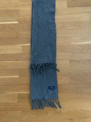 Grå halsduk från Polo Ralph Lauren - Mysig grå halsduk från Polo Ralph Lauren i mjuk ull. Klassisk design med fransar i ändarna och en liten broderad logga. Perfekt accessoar för kyliga dagar.