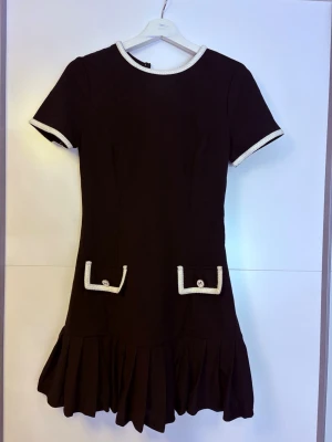 Svart kortklänning med volangkant - Supersöt svart kortklänning med vita detaljer vid ärmslut och halsringning. Klänningen har två dekorativa fickor framtill med vita kanter och guldknappar med rosett. Kjolen avslutas med en bred volangkant som ger extra rörelse. Perfekt för dig som gillar klassisk men lekfull stil.