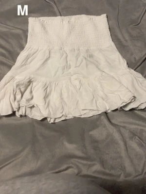 Vit volangkjol i bomull, storlek M - Supersöt vit kortkjol med bred smock i midjan och volang längst ner. Kjolen är gjord i mjuk bomull och har en luftig känsla som passar perfekt till varma dagar. Enkel att matcha med crop tops eller linnen för en chill look.