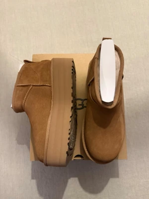 Bruna UGG  - Säljer ett par bruna UGG boots i mocka med rund tå och rejäl platåsula. Skorna har en klassisk UGG-look med mjukt foder på insidan och UGG-logga bak på hälen. Perfekta för höst och vinter när du vill ha både stil och komfort.