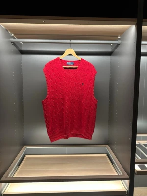 Röd stickad väst från Polo Ralph Lauren - Säljer en röd stickad väst från Polo Ralph Lauren i 100% pima bomull. Klassisk kabelstickad design med v-ringning och liten mörkblå logga broderad på bröstet. Perfekt att slänga över en skjorta för en preppy look.                                                              Längd:67cm.     Bredd:45cm
