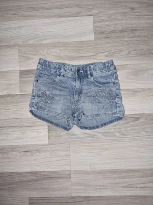 Ljusblå jeansshorts med stjärnor H&M - Snygga ljusblå jeansshorts från H&M med coola stjärndetaljer i nitar på både fram- och bakficka. Klassisk femficksmodell med knapp och dragkedja. Perfekta för sommaren och ger en avslappnad vibe.