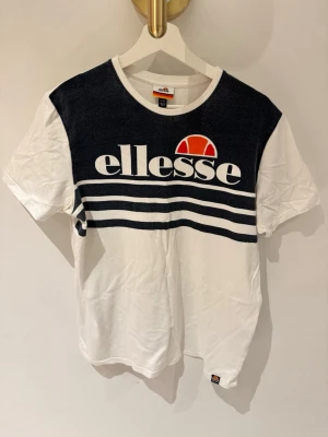 Vit och svart Ellesse t-shirt - Snygg t-shirt från Ellesse i vitt och svart med stor logga och orange detalj framtill. Klassisk rund hals och korta ärmar. Tillverkad i mjuk bomull och har två vita ränder över bröstet för en sportig vibe.