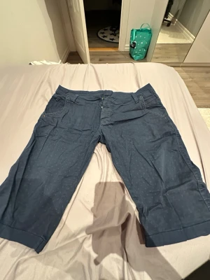 Blå caprishorts från ORJEAN, stl 34 - Säljer ett par blå caprishorts från ORJEAN  i storlek 34. Shortsen har diskret prickigt mönster, klassisk femficksdesign och knäppning med knappar. Materialet känns som bomull och de har en avslappnad passform som är perfekt för varmare dagar.