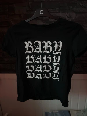 Svart t-shirt med BABY-tryck - Svart t-shirt med vitt BABY-tryck i gotisk stil på framsidan. Klassisk rund hals och korta ärmar. Tillverkad i mjukt bomullstyg som är skönt mot huden. Perfekt för dig som gillar statement-plagg och streetwear-vibe.