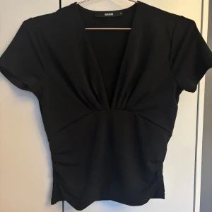 Svart v-ringad topp från Bik Bok - Säljer en svart topp från Bik Bok med v-ringning och korta ärmar. Toppen har en snygg drapering framtill och är tillverkad i ett mjukt, stretchigt material.