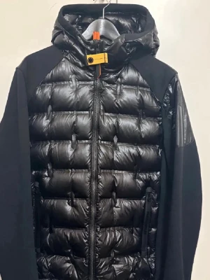 Svart pufferjacka från Parajumpers - Säljer en svart pufferjacka från Parajumpers med huva och quiltad design. Jackan har dragkedja framtill, två sidofickor med dragkedja och en gul detalj vid halsen. Materialet är en mix av polyester och dun, vilket gör den både varm och lätt. Perfekt för kalla dagar.