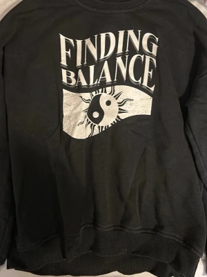 Svart sweatshirt Finding Balance 157 - Cool svart sweatshirt från 157 med stort vitt tryck på bröstet där det står 'Finding Balance' och en yin-yang-symbol med solstrålar. Tröjan har rund hals, ribbade muddar och är långärmad. Perfekt för dig som gillar statement-plagg.