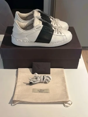 Valentino Garavani vita sneakers - Säljer ett par vita Valentino Garavani sneakers med svart bred rem över mitten.   Skorna har rund tå, snörning och coola nitar på hälen. Tillverkade i skinn och kommer med extra skosnören och dustbag. Perfekta för dig som vill ha en clean men lyxig look.