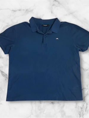 Ljusblå piké från J.Lindeberg XL - Snygg ljusblåblå pikétröja från J.Lindeberg med klassisk krage och korta ärmar. Tröjan har en liten vit logga broderad på bröstet och knappar vid halsen. Perfekt till jeans eller shorts för en clean look.