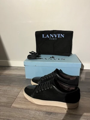 Svarta sneakers från Lanvin i mocka och lack - Säljer ett par svarta Lanvin sneakers med ovandel i mjuk mocka och detaljer i blankt kalvskinn. Skorna har vita gummisulor, rund tå och klassisk snörning. Kommer med originalkartong och extra skosnören. Riktigt stilrena och tidlösa sneakers från Lanvin Paris. Storlek 40-41