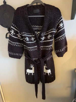 Brun stickad kofta med renar från Nümph - Mysig brun stickad kofta från Nümph med vita detaljer och renar. Koftan har knappar framtill, bälte i midjan och trekvartsärmar. Perfekt för kalla dagar och har ett klassiskt vintermönster som sticker ut.