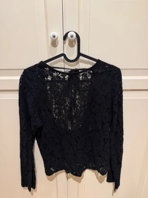 Svart spetsblus med knyt i ryggen - Säljer en svart blus i spets med lång ärm. Toppen är öppen i ryggen med knytband och spetsmönster. 