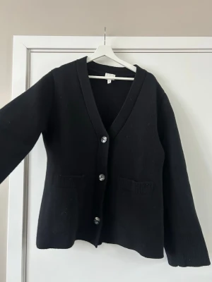 Svart kofta i ull med knappar - Klassisk svart kofta med v-ringning och stora knappar framtill. Två fickor fram och ribbade muddar vid ärmslut och fickor. Tillverkad i 98% ull och 1% elastan. Är inte oversized i modellen, går lite inåt vid midjan. 