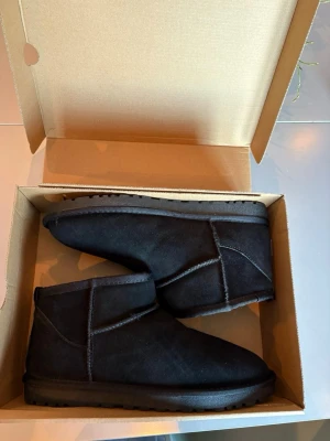 Svarta UGG boots i mocka storlek 45/44 - Säljer ett par klassiska svarta UGG boots aldrig använda i mjuk mocka med rund tå och platt sula. Skorna har UGG-logga bak på hälen och är fodrade för extra värme. Perfekta för kalla dagar och riktigt sköna att ha på sig.