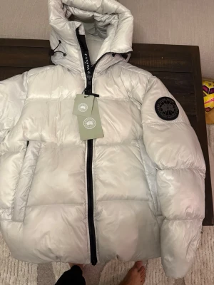 Ljusgrå  Canada Goose - Snygg ljusgrå pufferjacka från Canada Goose med stor huva, svarta detaljer och patch på ärmen. Jackan har dragkedja framtill, två stora fickor och justerbar snörning vid huvan. Insidan har svarta axelband med logga och meshfickor. Helt ny finns I flera storlekar.