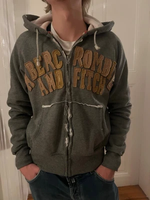 Abercrombie & Fitch zip hoodie – Medium - Snygg och klassisk Abercrombie & Fitch hoodie med dragkedja i grön/grå vintage-ton. Broderad logotyp framtill i bruna/beige nyanser ger en tidlös college-look. Mjuk och varm insida – perfekt för vardag, skola eller casual outfits. ✔ Original Abercrombie & Fitch ✔ Storlek: Medium ✔ Färg: grön/grå med beige insida ✔ Skick: Bra begagnat skick, normalt slitage (se bilder) ✔ Bekväm passform & hög kvalitet Passar perfekt till jeans, cargobyxor eller som lager under jacka. Svår att hitta i denna stil! 
