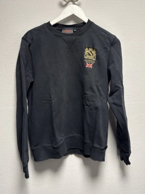Svart sweatshirt från Morris - Svart sweatshirt från Morris med broderad guldfärgad logga och brittisk flagga på bröstet. Tröjan har rund hals, ribbade muddar och är långärmad. Perfekt för dig som gillar klassisk och stilren streetwear. Skriv angående frågor!