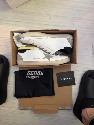 Golden Goose Superstar sneakers vit/grå - Golden Goose Superstar sneakers i vitt skinn med grå mockadetaljer och stjärna på sidan. Grå snörning, svart häl och slitna detaljer på sulan för en cool vintage-look. Kommer med originalpåse och certifikat.