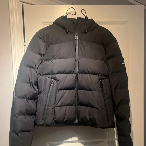 Svart pufferjacka från Superdry - Säljer en svart pufferjacka från Superdry med huva och dragkedja framtill. Jackan har två snedställda fickor med dragkedja och ett broderat Superdry-märke på ärmen. Perfekt för kalla dagar och riktigt snygg till streetwear.