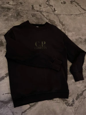 Svart sweatshirt från C.P. Company - Säljer en svart sweatshirt från C.P. Company med tryckt logga på bröstet. Tröjan har rund halsringning, långa ärmar och ribbade muddar vid ärmslut och nederkant. Perfekt för dig som gillar stilrena och klassiska plagg. M/L