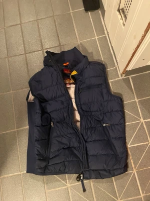 Mörkblå dunväst från Parajumpers - Säljer en mörkblå dunväst från Parajumpers med hög krage, gul detalj vid halsen och flera dragkedjeförsedda fickor. Västen har quiltad design och patch på ryggen. Perfekt för lager-på-lager och riktigt snygg till jeans.