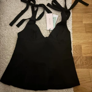 Svart satinlinnetopp med knytband - Supersnygg svart linnetopp i satin med breda axelband som knyts på axlarna. Toppen har en v-ringad skärning och lätt utsvängd nederdel för en feminin siluett. Perfekt att styla med jeans eller kjol för en trendig look. Aldrig använd + prislappen kvar, säljer då den inte passar❣️