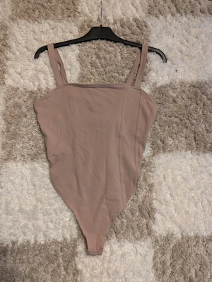 Beige body med breda axelband - Säljer en enkel och stilren beige body med breda axelband och raka sömmar framtill. Bodyn har en mjuk känsla och är tillverkad i stretchigt bomullsmaterial. Perfekt att styla med jeans eller kjol för en clean look.