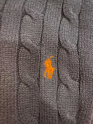 Mörkblå kabelstickad tröja Polo Ralph Lauren - Kabelstickad mörkblå tröja från Polo Ralph Lauren i storlek L. Klassisk modell med rund hals och orange broderad logga på bröstet. Tillverkad i mjuk bomull och perfekt för lager på lager under höst och vinter.
