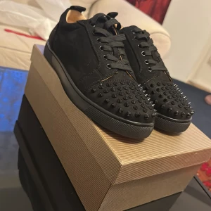 Christian Louboutin svarta sneakers med nitar - Säljer ett par svarta sneakers från Christian Louboutin med ikonisk röd sula och svarta nitar över tån. Skorna har snörning, rund tå och är tillverkade i mocka med detaljer i skinn. Perfekta för dig som vill sticka ut med edgy design och lyxig känsla.