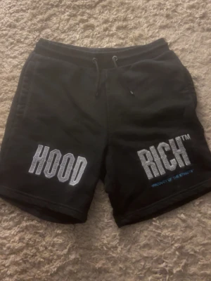 Svarta Hoodrich shorts med tryck - Svarta shorts från Hoodrich med stora vita och gråa tryck på framsidan där det står HOODoch RICH. De har snörning i midjan och är gjorda i mjukt bomullsmaterial som gör shortsen skön att använda. Perfekta för chill dagar eller streetwear-stil. Använt runt 3 gånger så det är inget fel med dom.