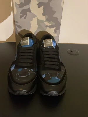 Valentino rockrunners  - Säljer ett par Valentino Garavani sneakers i svart med blå och grå camouflage-detaljer. Skorna har snörning, rund tå och platt sula. Märkeslogga på plösen och coolt mönster på ovandelen. Perfekta för dig som vill sticka ut med exklusiva sneakers.