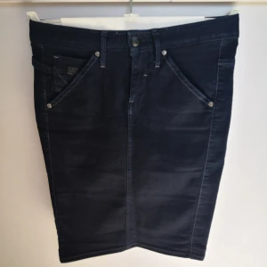Mörkblå denim kjol från G-Star RAW - Mörkblå denim kjol från G-Star RAW med klassiska femficksdetaljer, bälteshällor och knappstängning. Rak passform med strech och bakfickor med dekorativa sömmar. Kjolen är 50 cm lång, och är i storlek 26. 