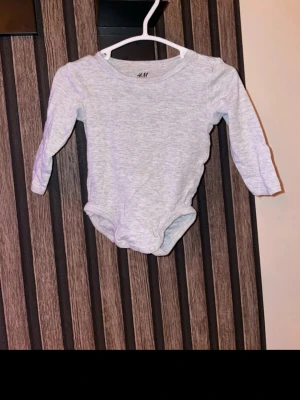 Grå långärmad body från H&M storlek 62 - En enkel och mjuk grå långärmad body från H&M. Tillverkad i bomull med rund hals och tryckknappar i grenen för enkel påklädning. Perfekt basplagg till garderoben. Storlek 62