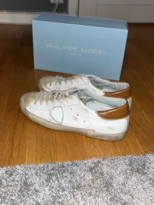 Vita Philippe Model sneakers i skinn - Säljer ett par vita Philippe Model sneakers i skinn med beige detaljer och bruna hälpartier. Klassisk låg modell med snörning och rund tå. Sulan har tydlig Philippe Model-logga undertill. Perfekta för dig som gillar stilrena och tidlösa sneakers. Har andvänt dom ett få tal gånger under 1 vecka inget mer.