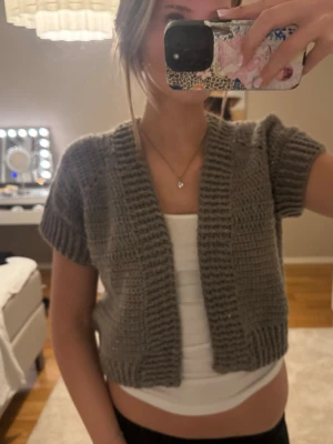 Grå stickad kortärmad kofta - Säljer en handstickad grå kofta med korta ärmar och öppen framtill. Koftan är gjord i ett tjockare garn med små färgade noppdetaljer och har bred ribbad kant vid ärmslut och nederkant. Perfekt att slänga över ett linne för en chill look.
