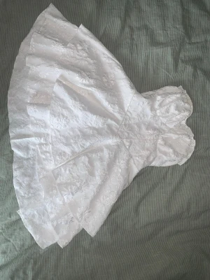 Vit axelbandslös klänning med blommigt mönster - Charmig vit axelbandslös klänning som formar midjan med ett vackert blommigt mönster. Klänningen har en volangdetalj Den har en kort längd och en elegant passform som framhäver figuren. Bust:74-100 cm, Waist Size:67-96 cm, Length:64 cm