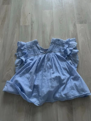 Ljusblå volangblus i bomull - Passar s/m Supersöt ljusblå blus med volangdetaljer på ärmarna och smock upptill. Blusen har en luftig passform och är gjord i mjuk bomull, perfekt för varma dagar. V-ringad hals och romantisk känsla. 