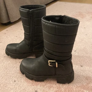 Svarta boots med spänne och dragkedja - Tuffa svarta boots i skinnimitation med grov sula, guldfärgat spänne och dragkedja på sidan. Insidan är fodrad för extra värme och skaftet är quiltat för en cool look. Perfekta för höst och vinter när du vill ha både stil och funktion. Har några lösa trådar och är liiiite sliten, men ger mest en cool och grunge vibe 😎Har också lite smuts på sulan och på remmarna men det går att tvätta bort! 💛💛💛