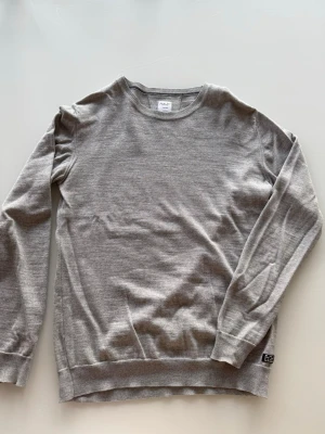 Grå stickad tröja från Seven Seas - Snygg grå stickad tröja från Seven Seas Copenhagen i storlek medium. Tröjan har rund hals, ribbade muddar och är långärmad. Perfekt för dig som gillar enkel och stilren design med mjukt material.