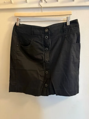 Svart kortkjol med knappar framtill - Säljer en svart kortkjol med raka linjer och knappar längs hela framsidan. Kjolen har fickor både fram och bak samt hällor för bälte. Materialet känns som bomull och modellen är klassisk och enkel att matcha.