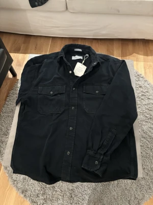 Svart overshirt från GANT - Säljer en svart overshirt från GANT i oversized fit. Skjortjackan har två bröstfickor med knappar, klassisk krage och tryckknappar framtill. Materialet är mjukt och känns slitstarkt, perfekt för lager på lager. Enkel och stilren design. (Lite åt de mörkblåa hållet) 