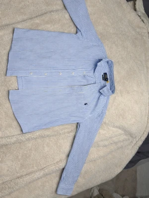  Polo Ralph Lauren - Säljer en skjorta från Polo Ralph Lauren i blåvitt randigt mönster. Skjortan har långa ärmar, knappar framtill och en broderad logga på bröstet. Tillverkad i mjuk bomull och har en snygg krage. Liten i storleken och de är barn modell. Helt ny de är därför priset är som de är kostar 1095 igentligen.