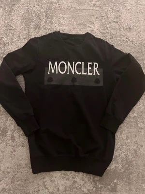 Svart Moncler sweatshirt med logga - Säljer en svart sweatshirt från Moncler med stor vit logga framtill och tre diskreta svarta emblem under. Tröjan har rund halsringning, långa ärmar och ribbade muddar vid ärmslut och nederkant. Perfekt för dig som gillar stilrena och exklusiva plagg.