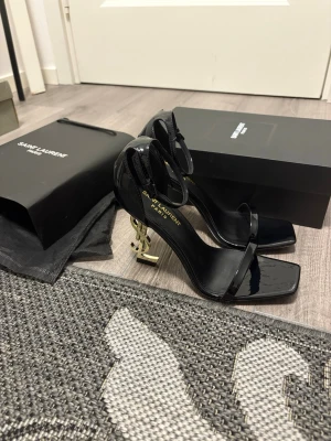 Saint Laurent svarta klackar med YSL-häl - Säljer ett par svarta Saint Laurent klackar i lackat skinn med ikonisk guld YSL-logga som klack. Skorna har fyrkantig tå, smal rem över tårna och justerbar ankelrem. Riktigt statement piece för dig som vill sticka ut.