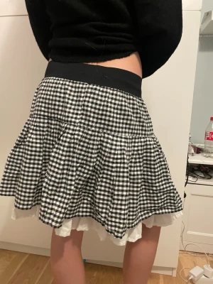 Rutig svartvit kortkjol med volang - Supersöt kortkjol i svartvitt rutigt mönster med bred svart midjeresår. Kjolen har volangig form och ett vitt spetskantat underlager som sticker fram nedtill. Perfekt för dig som gillar en lekfull och trendig stil.