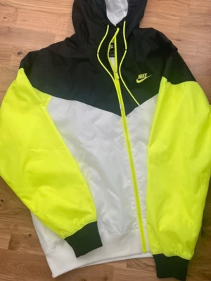 Nike vindjacka i svart, vit och neon - Nike vindjacka med huva i svart, vitt och neon-gult. Jackan har dragkedja framtill, elastiska muddar och meshfoder för extra komfort. Snygg kontrast mellan färgerna och klassisk Nike-logga på bröstet. Perfekt för dig som vill sticka ut. Ni får gärna diskutera pris