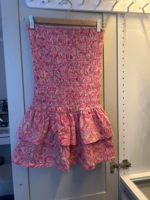 Rosa kortklänning från Gina Tricot - Supersöt kortklänning från Gina Tricot i rosa och ljusgult med smock upptill och dubbla volanglager nertill. Klänningen har ett blommigt mönster och är ärmlös, perfekt för varma dagar. Materialet känns lätt och somrigt. Perfekt för semester🩷🩷
